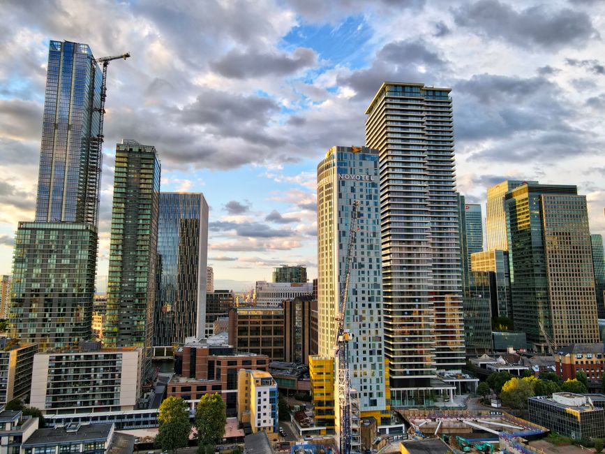 High-resolution photograph of a modern cityscape featuring several tall skyscrapers with reflective glass facades. The distinctive building on the left has a lattice-like exterior design, while the ce