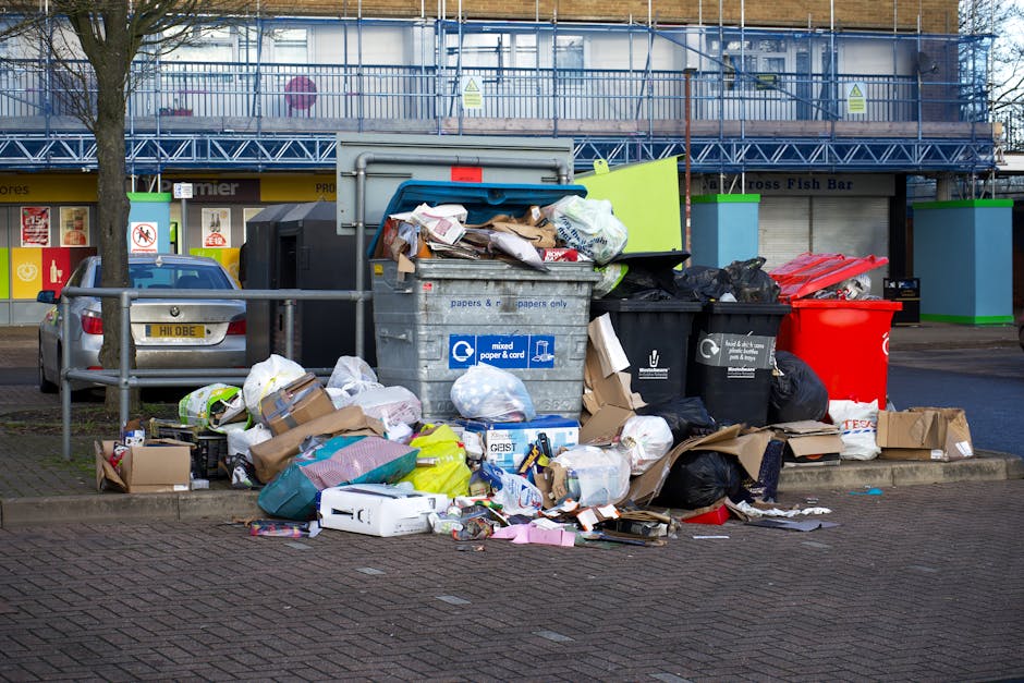 A row of four large white wheelie bins with closed lids, positioned on a paved surface outdoors, adjacent to a dark metal waste container. The bins are labeled 'WASTE ONLY' and are situated near a woo