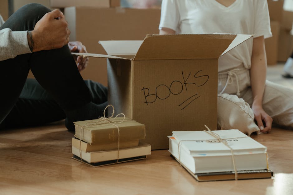Inside a home during a packing and moving process, a person is visible sitting on a wooden floor, partially out of the frame, holding a long strip of packing tape or strap. In front of them, there are