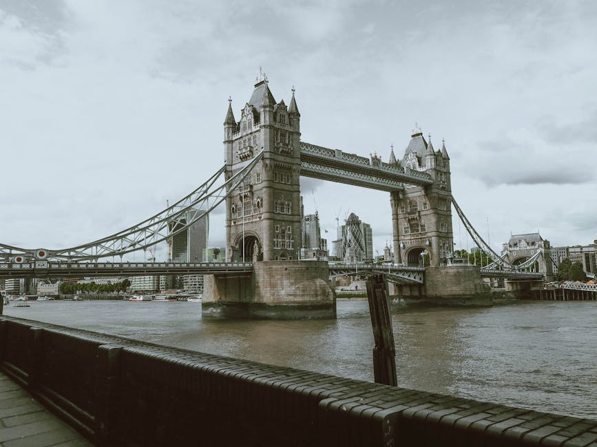 A high-angle view of Tower Bridge spanning the River Thames in London, featuring its iconic two towers with pointed spires and ornate stonework. The bridge's bascule and suspension components are visi