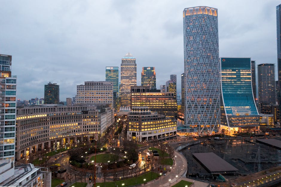 Photograph of the London skyline along the river Thames during the evening, with a focus on a variety of modern high-rise office buildings and residential apartments. The buildings are illuminated wit