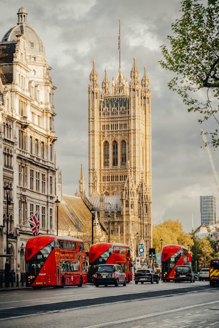 A panoramic view of the Palace of Westminster in London during late afternoon with clear skies. The historic Gothic-style building, featuring multiple spires and ornate detailing, is illuminated by wa